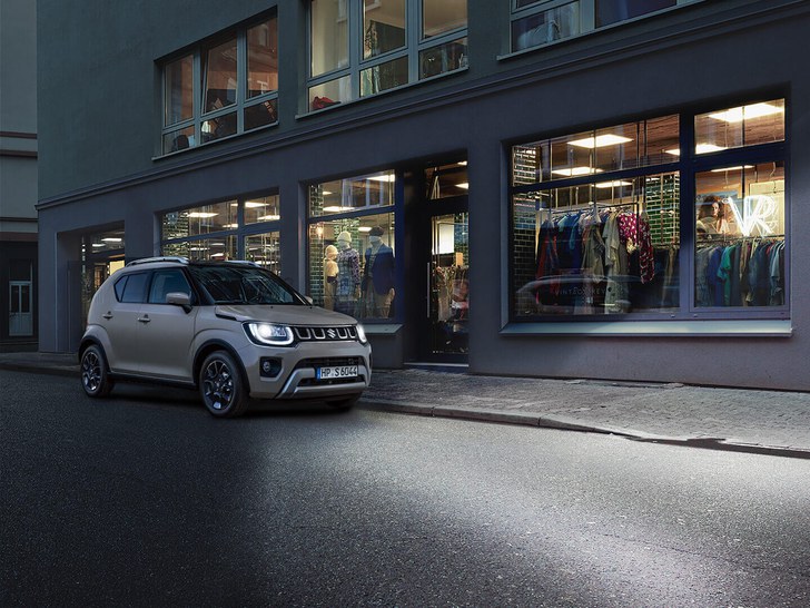 Ein Mann geht von einem Suzuki Ignis Hybrid in Caravan Ivory Pearl Metallic weg, welcher vor einem Modegeschäft geparkt ist.