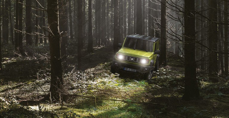 Suzuki Jimny Hybrid in Kinetic Yellow in einem nebeligen Wald, mit eingeschaltetem Licht.