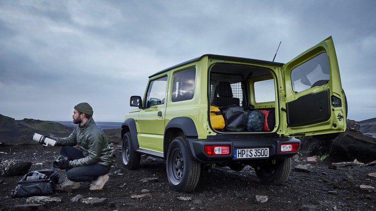 Suzuki Jimny Hybrid in Kinetic Yellow mit geöffnetem Gepäckraum, daneben ist ein Mann in der Hocke und schaut in die Ferne